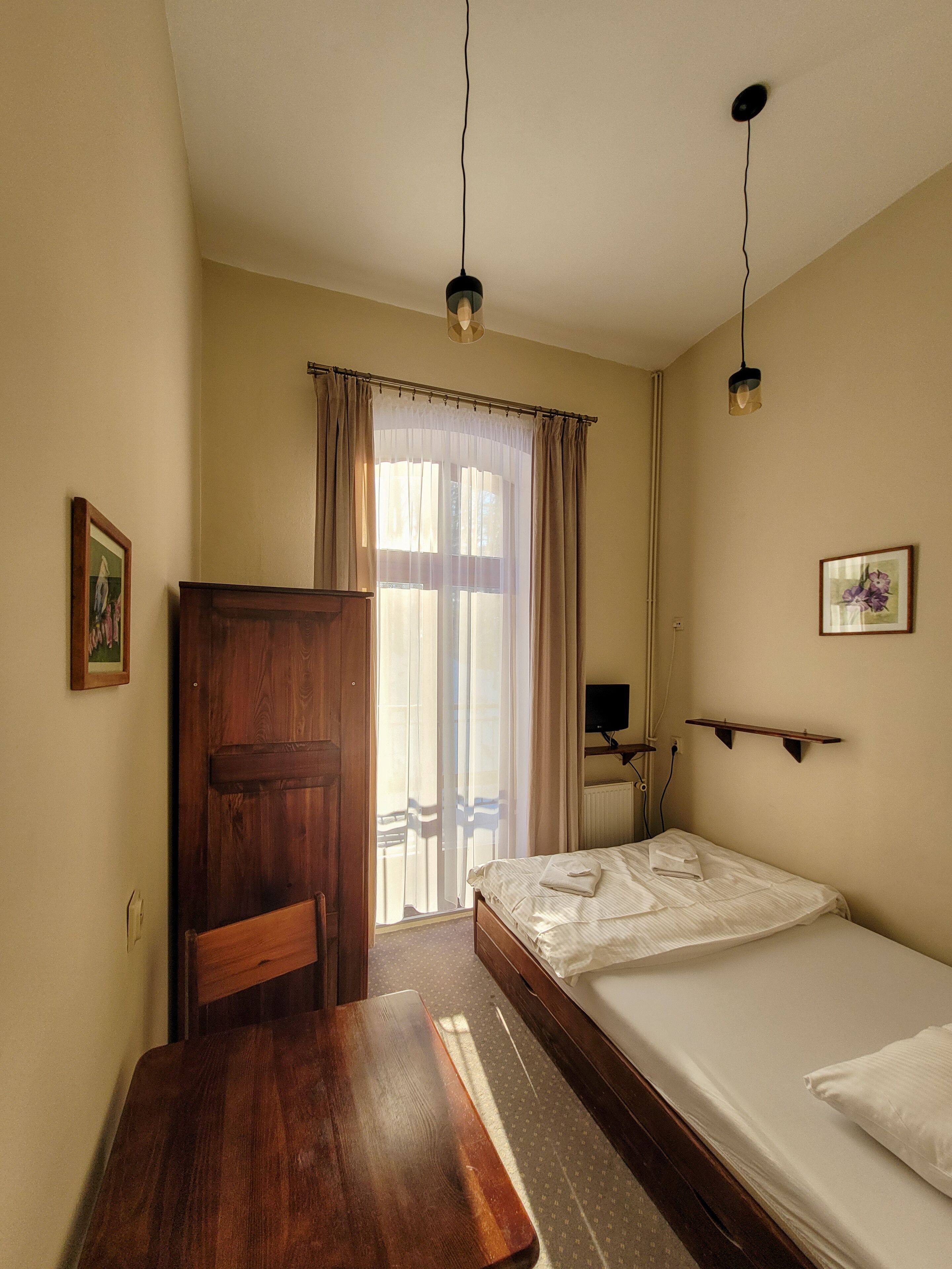 A room with a wooden desk, a bed, a chair, a window with curtains, and a wall-mounted shelf.