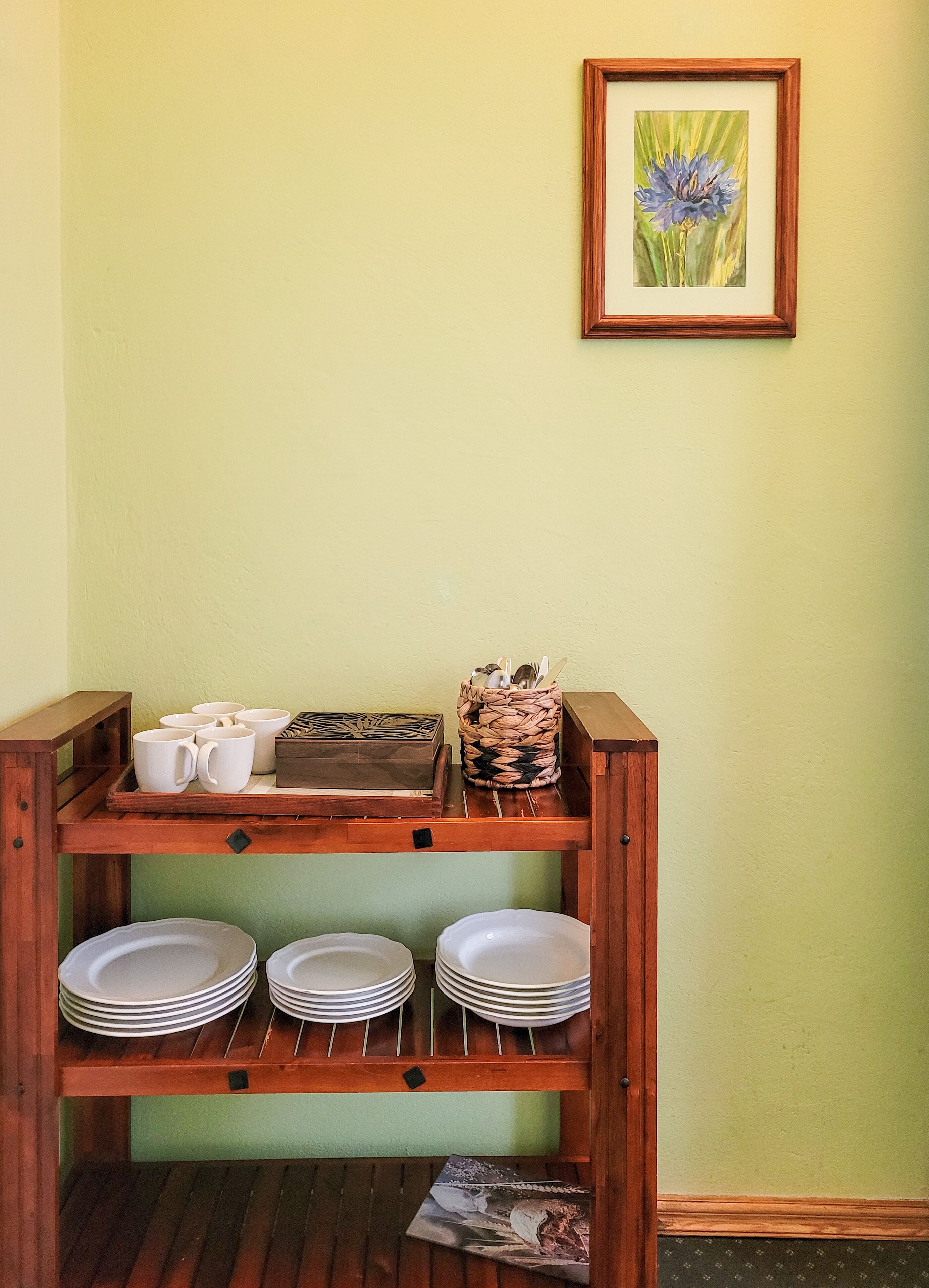 A wooden buffet stand with white plates and cups, a woven basket, and a framed picture of a flower on a green wall.