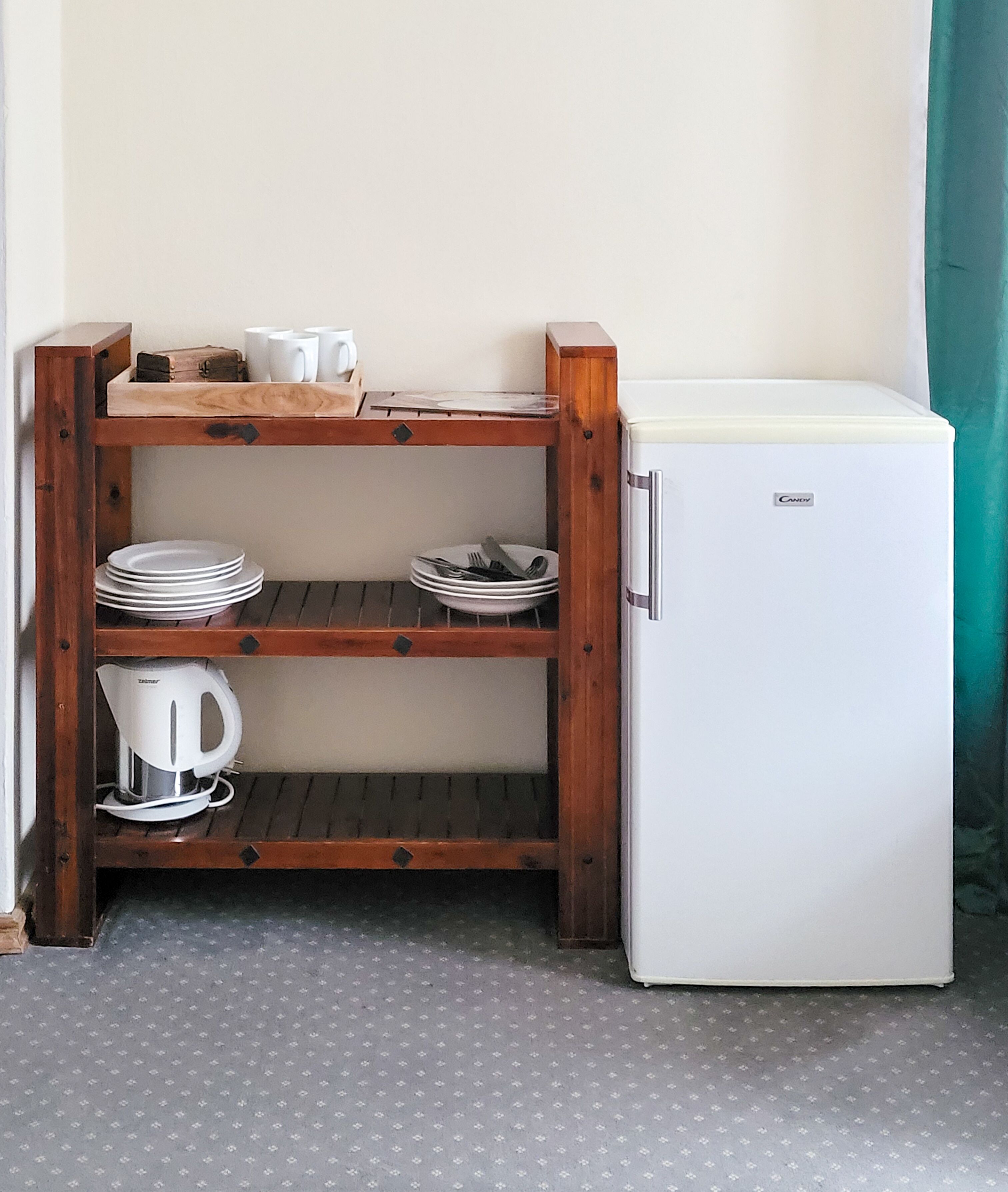 A compact kitchenette with a white refrigerator, wooden shelves holding plates and a kettle, and a small wooden table with cups.