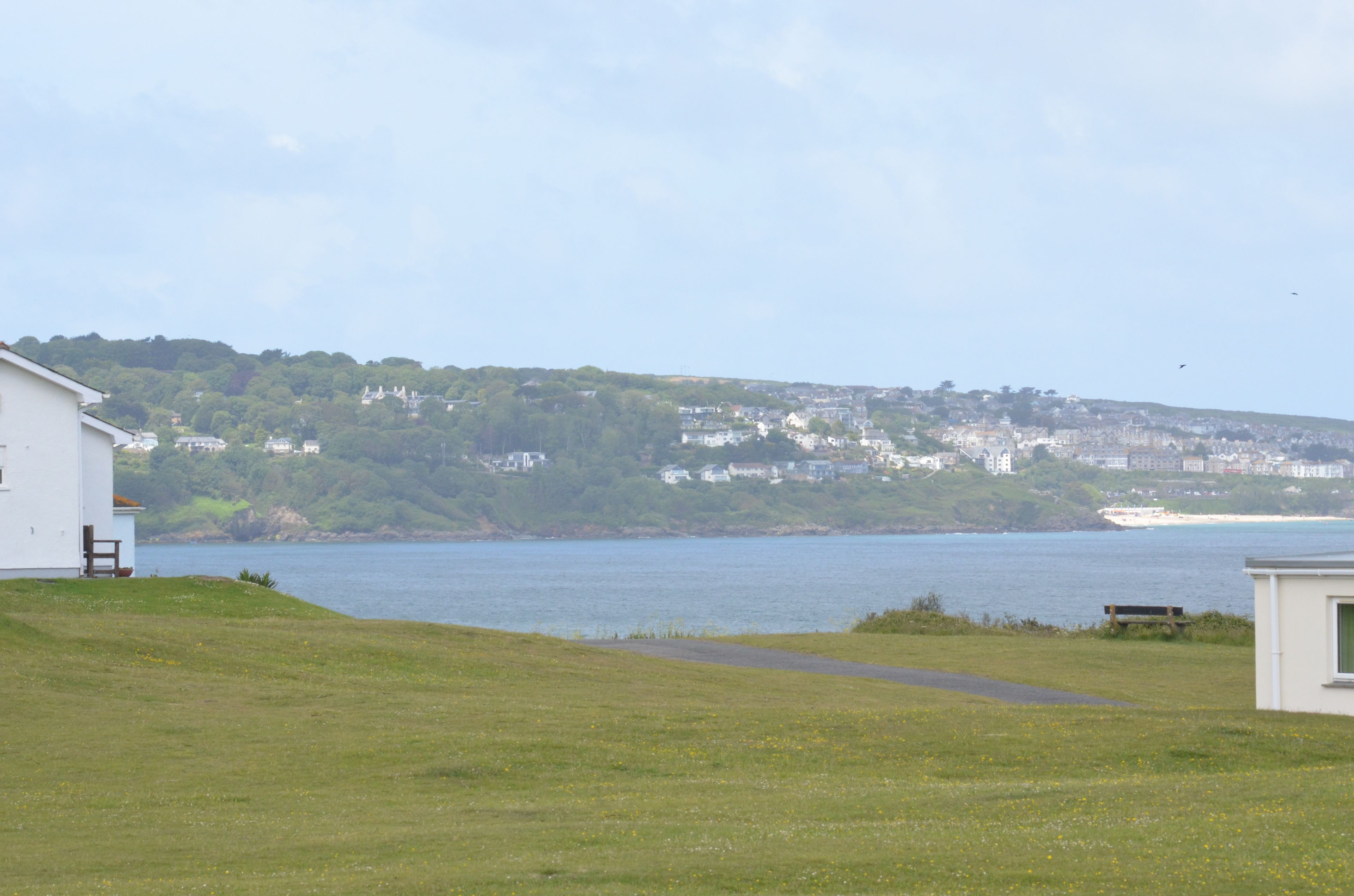 A coastal view with a grassy area, a white building, and a hill with houses in the background.