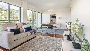 A modern living room with a sofa, a dining table, and a large window offering a view of buildings and greenery. - Monumental Apartment - By Wehost (Funchal)
