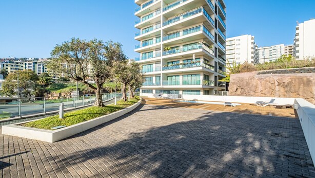 Un edifici modern de diversos pisos amb balcons, un passeig pavimentat i una zona enjardinada amb arbres.