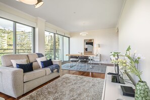 A modern living room with a sofa, a dining table, and a large window offering a view of buildings and greenery.