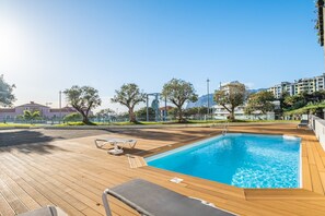Une piscine dotée d’une terrasse en bois et de chaises longues, entourée d’arbres et de bâtiments.