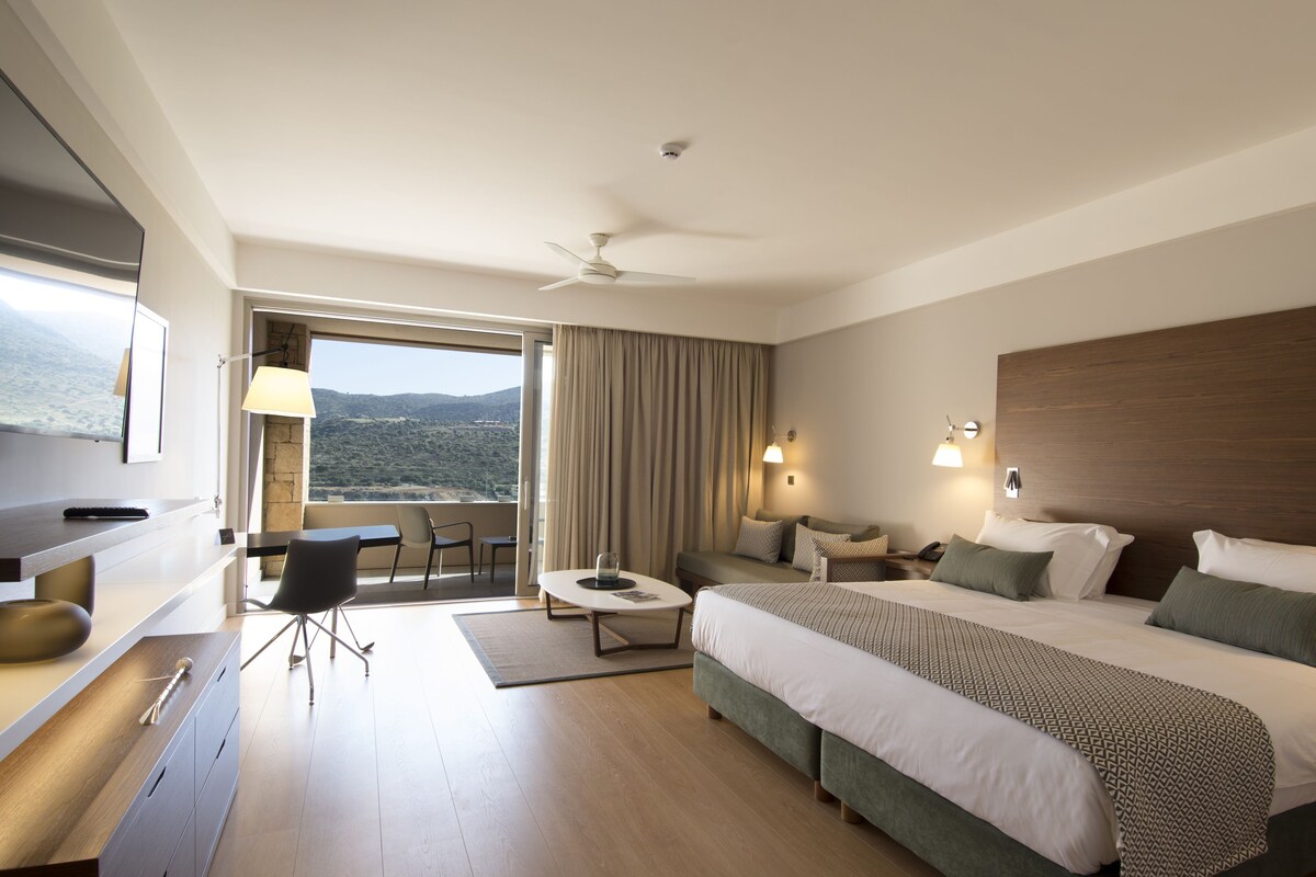 Habitación de hotel con cama, escritorio, silla, mesa, lámpara, ventilador de techo y vistas al exterior.