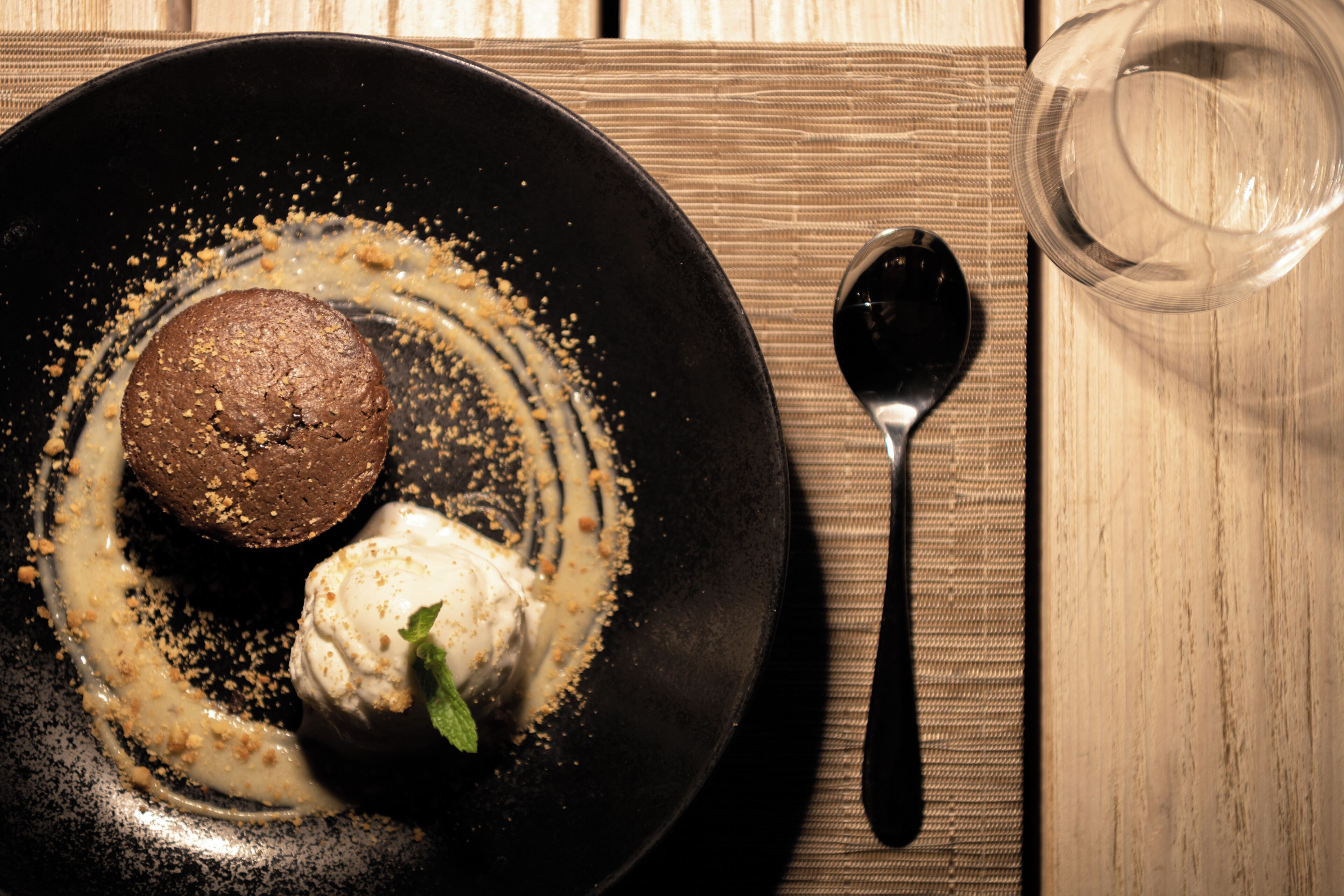 Un plat de postre amb un brownie i gelat, una cullera i un got d'aigua sobre una taula de fusta.