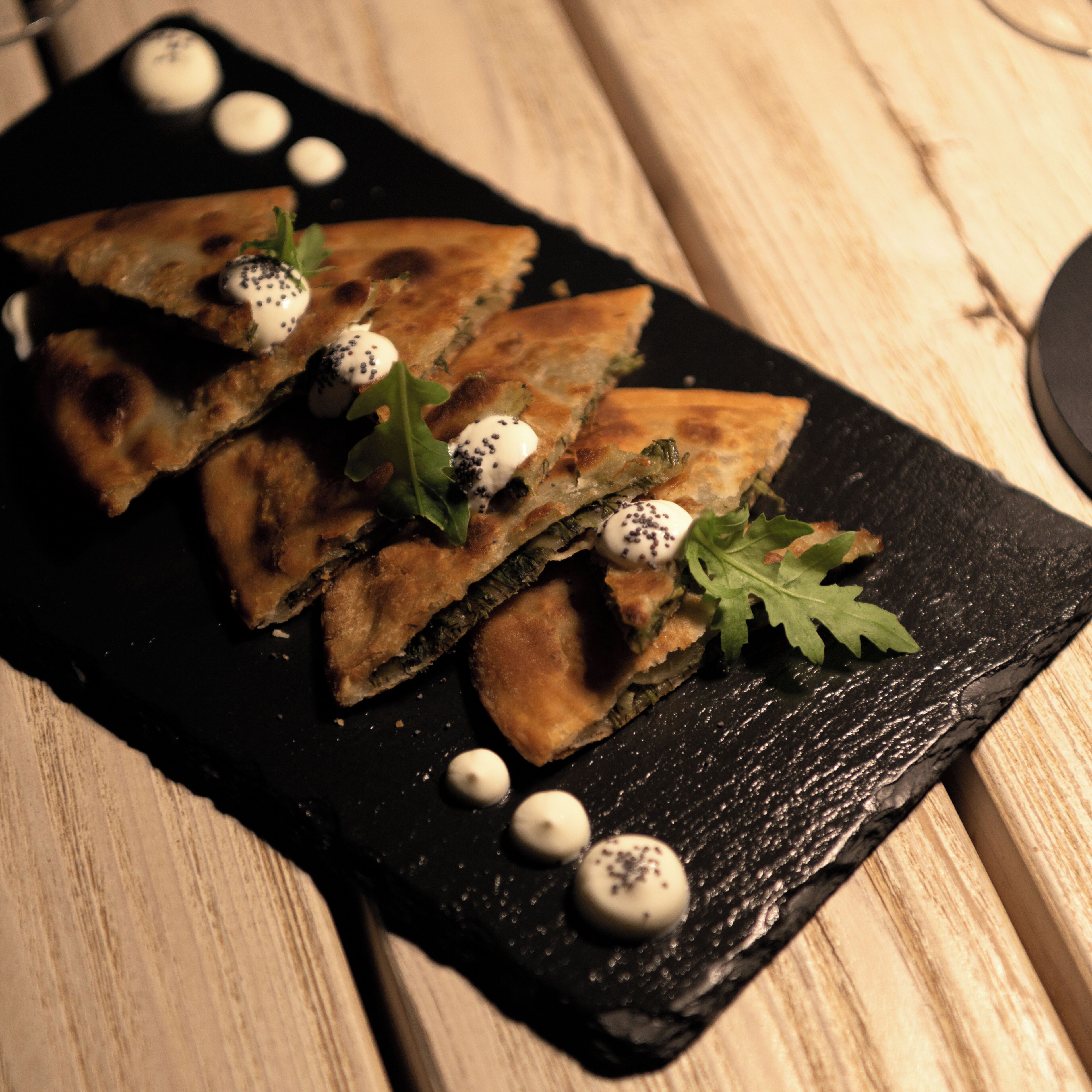 A black slate serving board with triangular food items, garnished with greens and white spheres, on a wooden table.