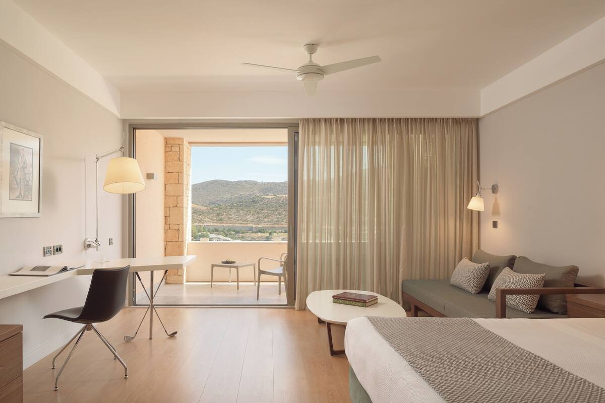 A hotel room with a bed, a desk, a chair, a small table, and a view of the outdoors.