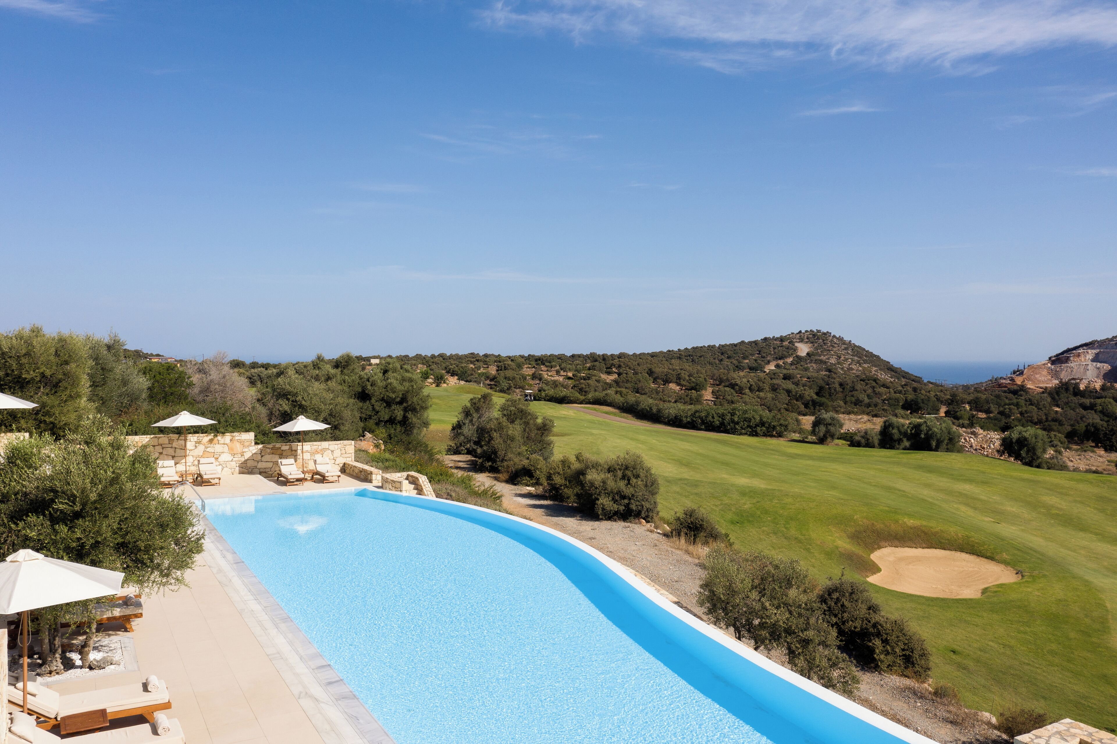 A swimming pool with lounge chairs and umbrellas, a golf course in the background, and a clear blue sky.
