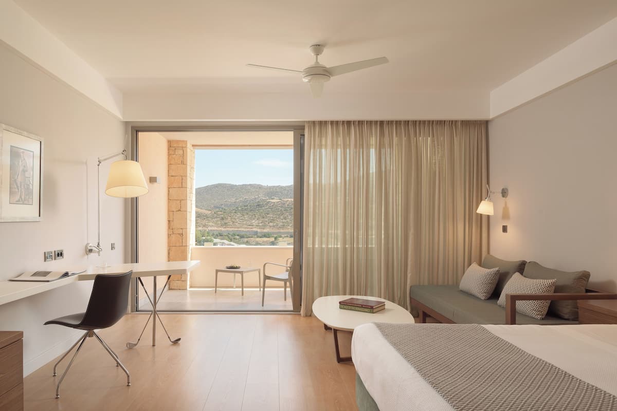 A hotel room with a bed, a desk, a chair, a small table, and a view of the outdoors.