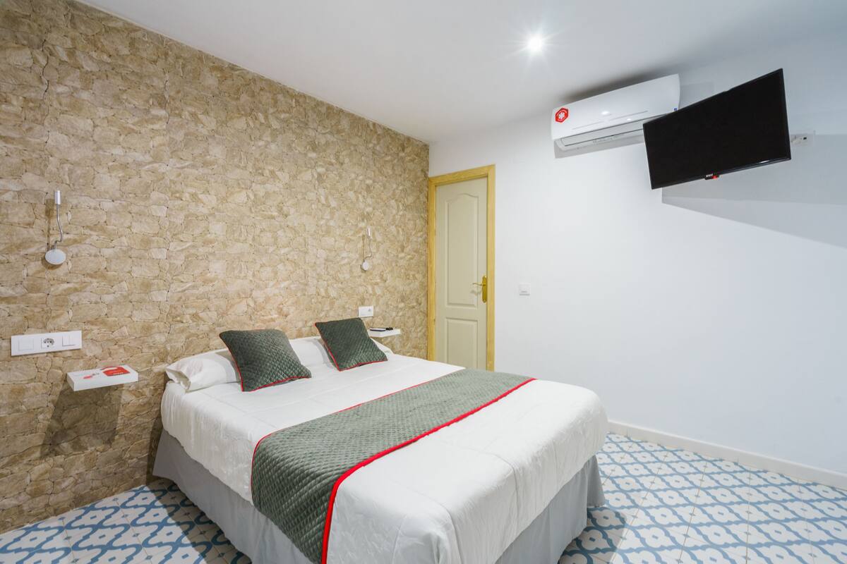 Un dormitorio con pared de piedra, cama con ropa de cama verde y blanca, televisor montado y baldosas de suelo estampadas.