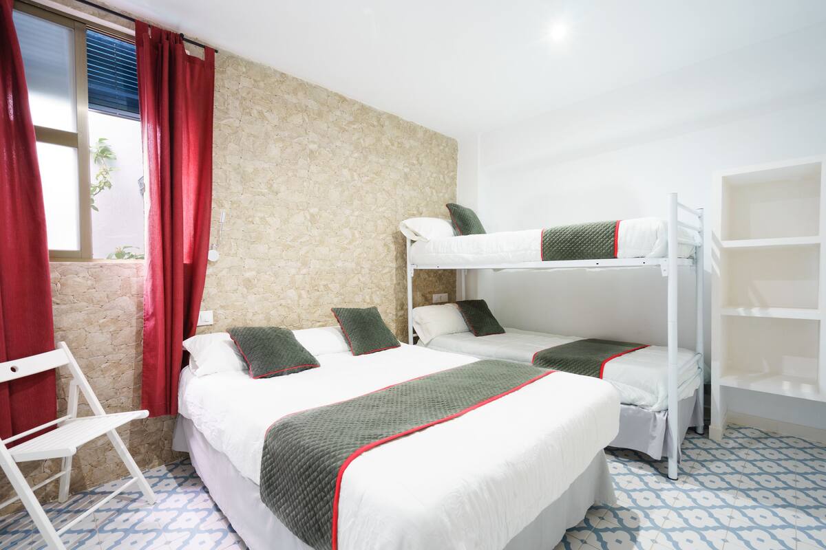 Habitación de hotel con literas, una silla blanca y una ventana con cortinas rojas.