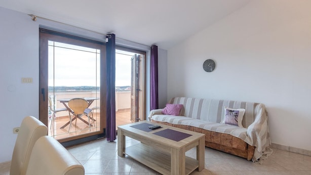 Un salon avec un canapé, une table basse et un fauteuil. Il y a une fenêtre avec des rideaux, une horloge au mur et une vue sur l’extérieur.