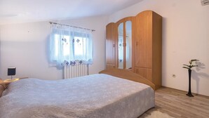 Une chambre à coucher comprenant un lit, une armoire en bois, une fenêtre avec des rideaux légers, une table de chevet et une plante en pot.