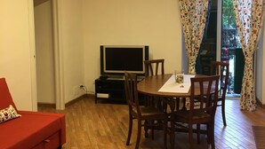 Un salon avec un canapé rouge, une table à manger en bois avec des chaises, un téléviseur et un balcon agrémenté de rideaux à motifs.