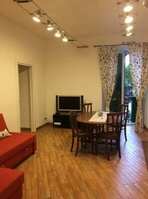 A room with a red sofa, a dining table with chairs, a TV, and a window with floral curtains.