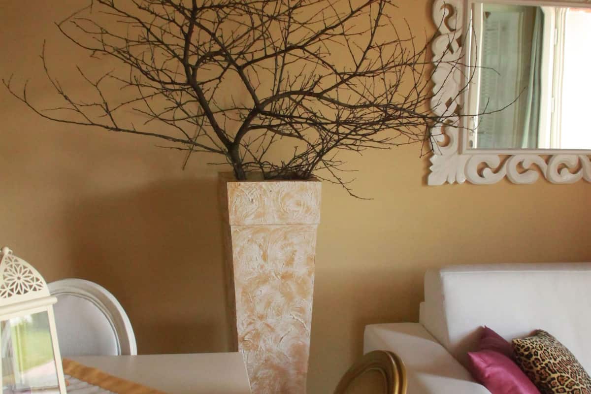 A room with a white sofa, a decorative vase with branches, a mirror with ornate frame, and a table with a patterned tablecloth.