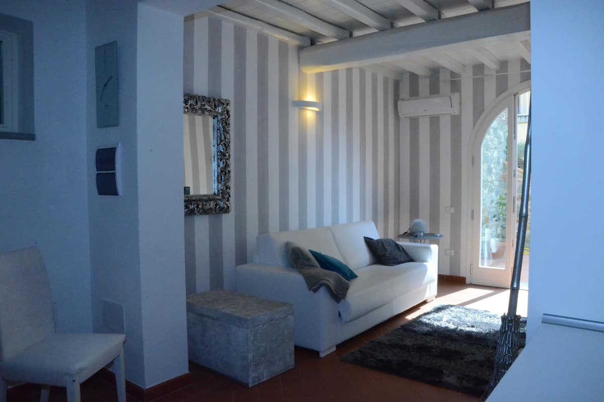 A room with a white sofa, a small ottoman, a chair, a mirror, and a window with a view of greenery.
