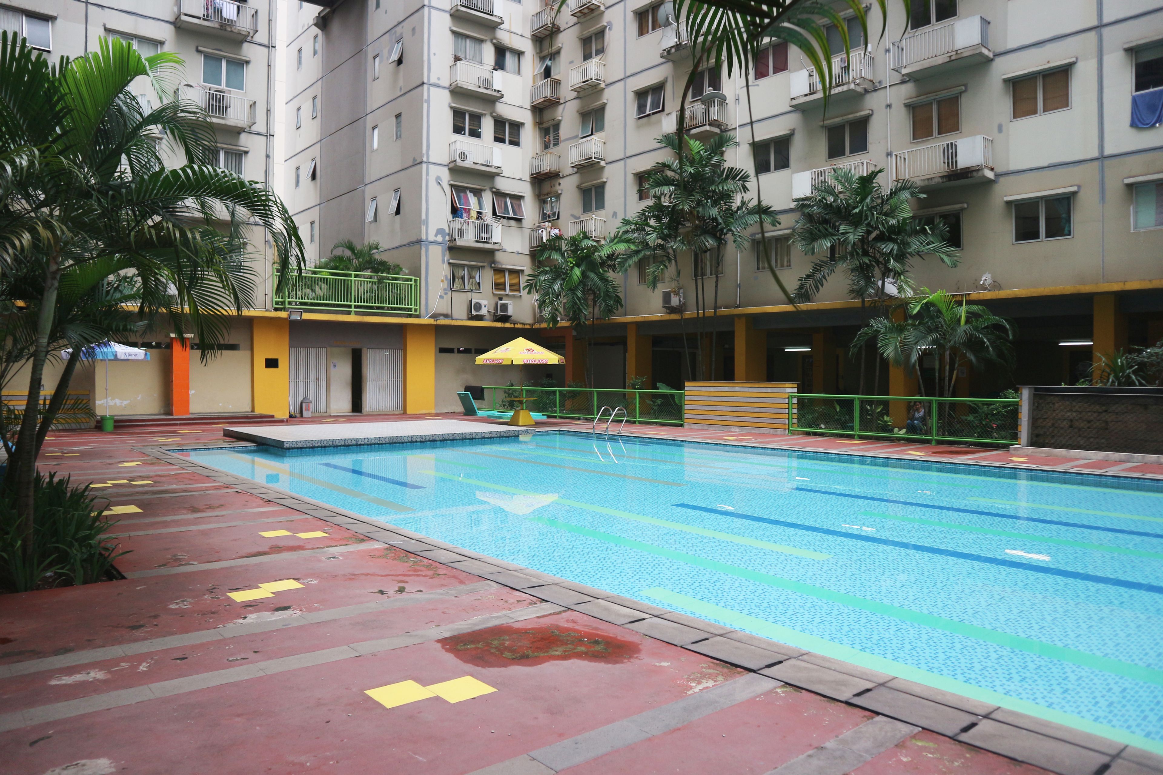 A swimming pool surrounded by a multi-story residential building with balconies, palm trees, and a yellow and red tiled walkway.