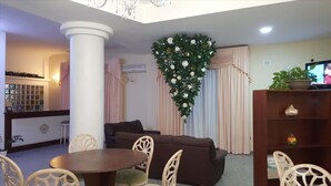 Een woonkamer met een bruine bank, een ronde houten tafel en witte stoelen. Er is een grote witte kolom, een decoratieve wand met een plant, en een televisie op een houten dressoir.