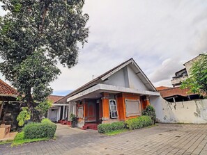 A two-story building with a tiled roof, a small porch, and a garden area with a large tree and shrubs.