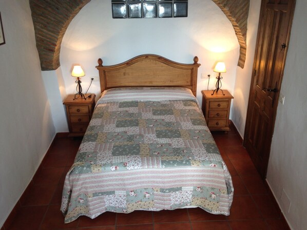 Un dormitorio con cabecera de madera, dos mesitas de noche con lámparas, colcha estampada y suelo de baldosas.