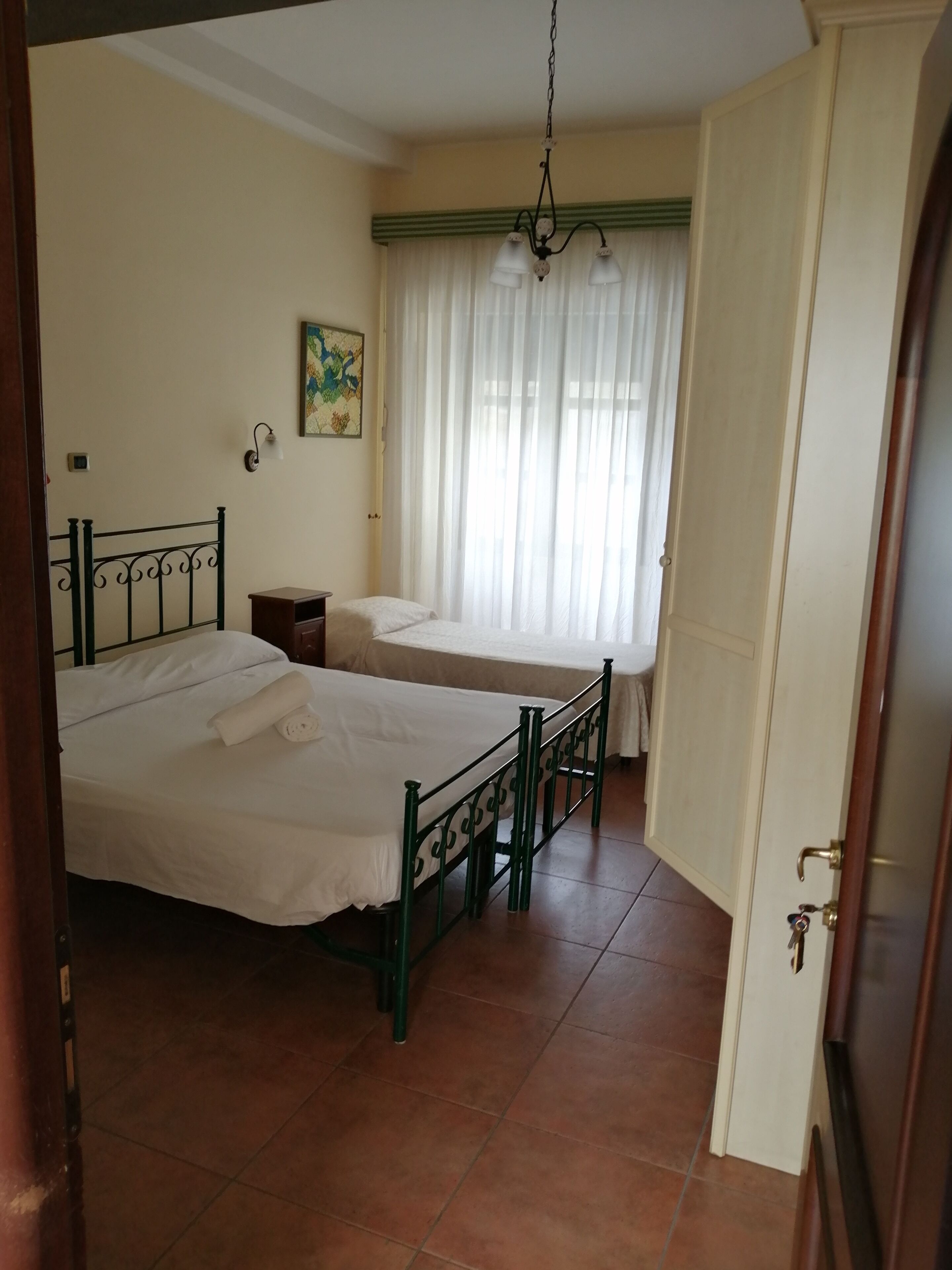 Une chambre avec deux lits simples, une petite table de chevet, une lampe fixée au mur et une fenêtre avec des rideaux légers.