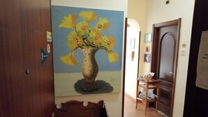 Um corredor com um banco de madeira, um quadro com um vaso e flores, e uma porta com uma placa.