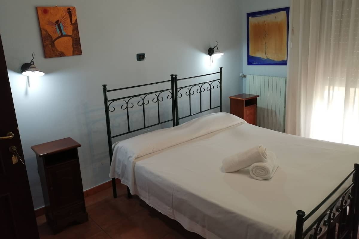 A bedroom with a metal bed frame, a bedside table, a wall-mounted lamp, and a painting.
