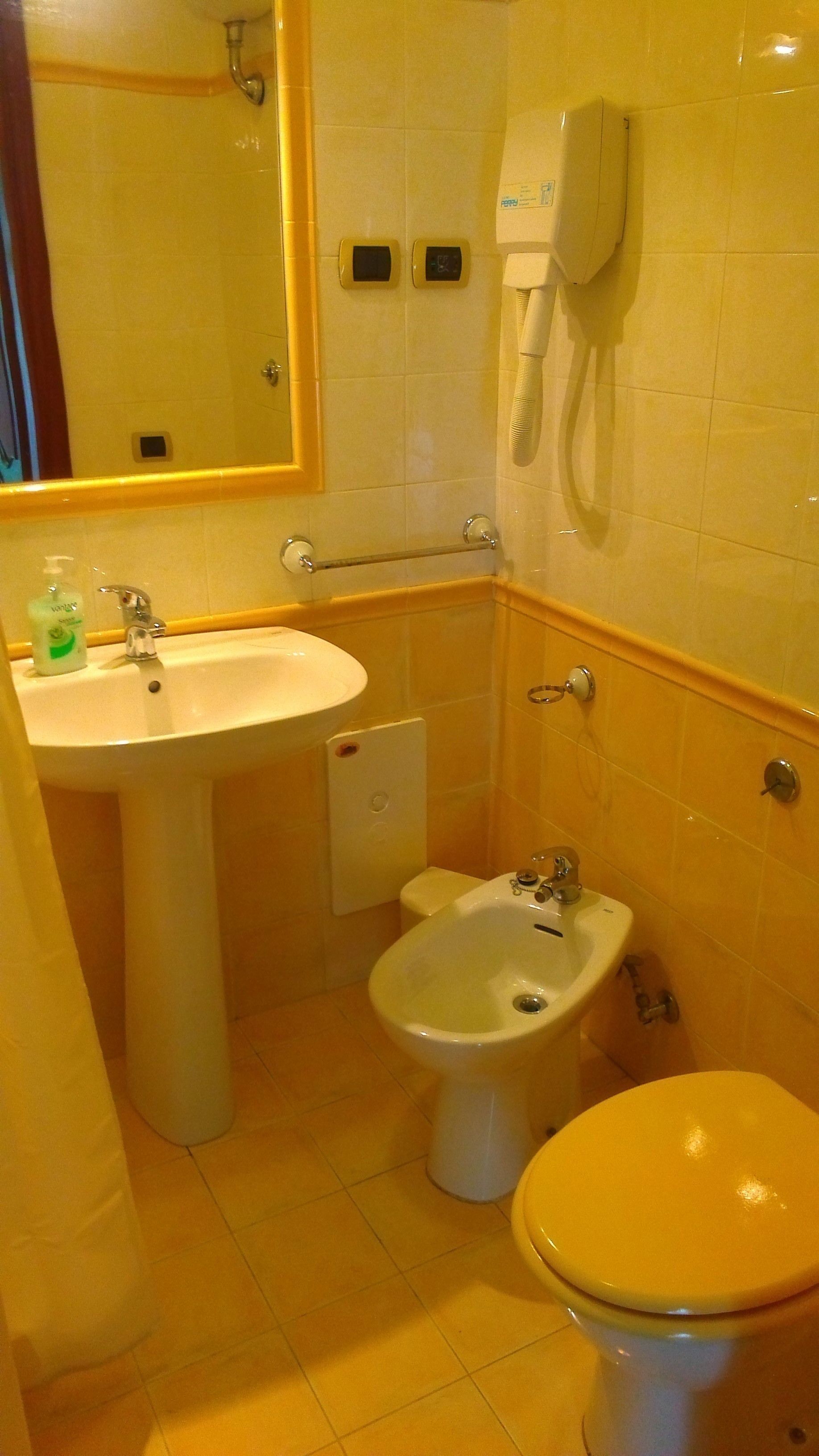 Une salle de bain équipée d’un lavabo, d’un bidet, de toilettes et d’un miroir. Un sèche-mains est fixé au mur et un distributeur de savon se trouve sur le lavabo.