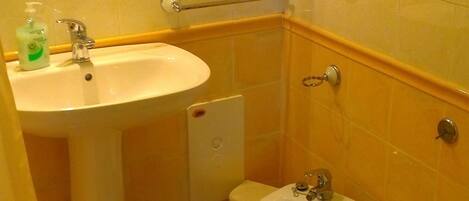 Un baño con lavamanos, bidé, inodoro y un espejo. Hay un secamanos en la pared y un dispensador de jabón en el lavamanos.