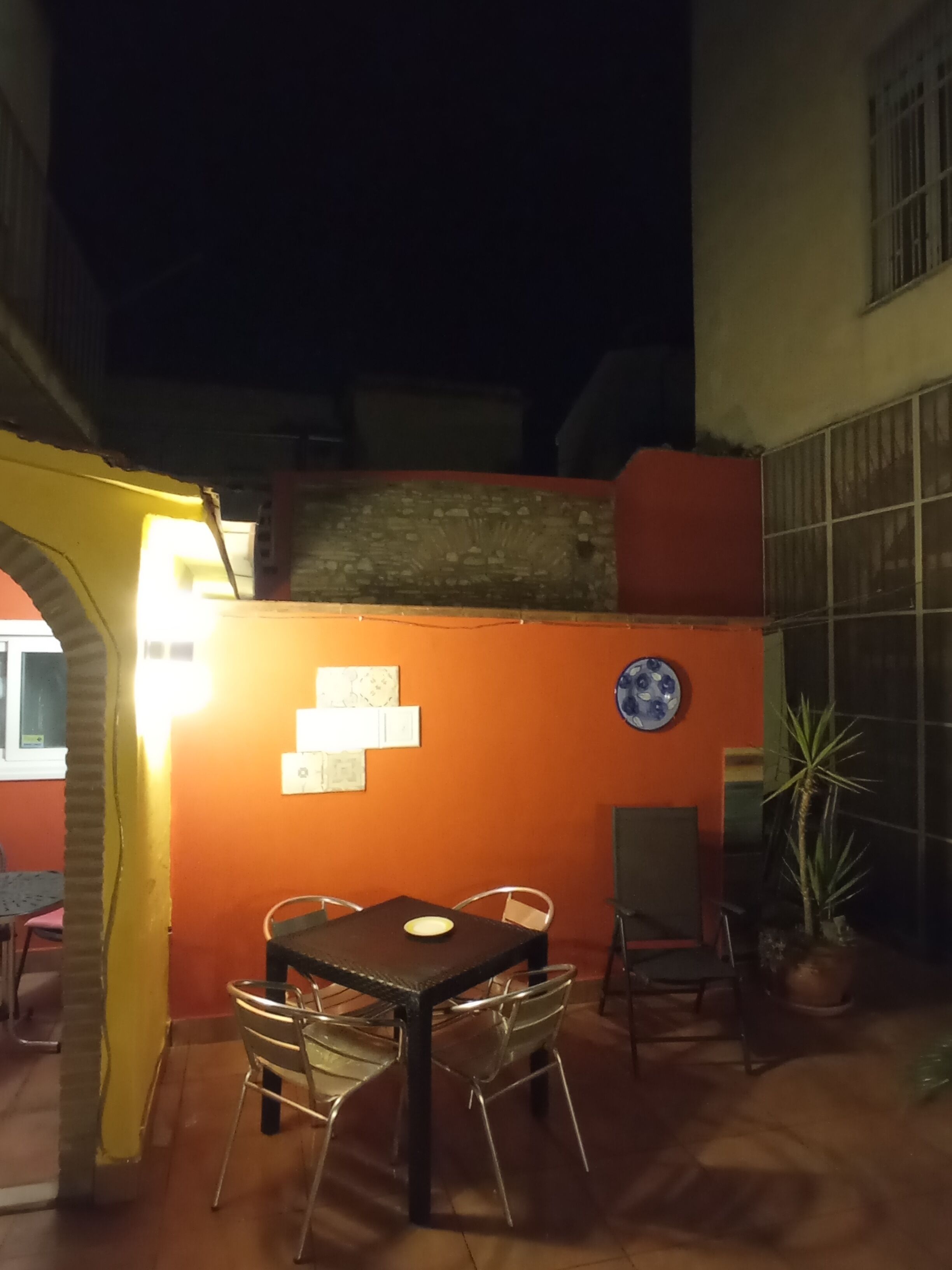Un patio al aire libre con una mesa y sillas, una planta en maceta, y una pared con un plato decorativo y una ventana.