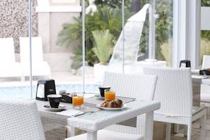 Ein Frühstücksbuffet mit weißen Korbstühlen und -tischen, einer Kaffeemaschine sowie einem Teller mit Croissants und Orangensaft.