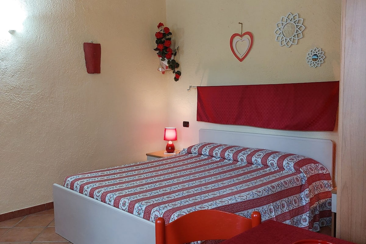 A bedroom with a bed, a nightstand, a red lamp, a red bucket, and a red tablecloth.