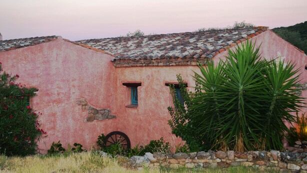 Un bâtiment rustique à la façade rose, doté d’un toit en tuiles et d’une roue placée devant.