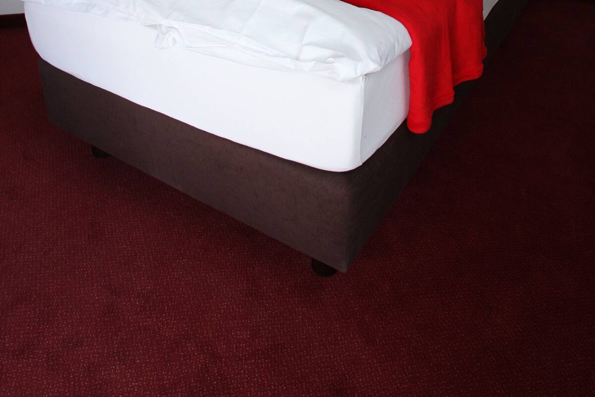 Ein ordentlich bezogenes Bett mit weißer und roter Bettwäsche, ein dunkles Kopfteil und ein rot ausgelegter Teppichboden.