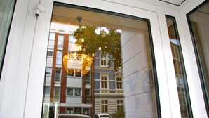 A view through a window of a building with a chandelier, a reflection of a tree, and a reflection of a building with multiple windows. - Hotel Gemini (Düsseldorf)