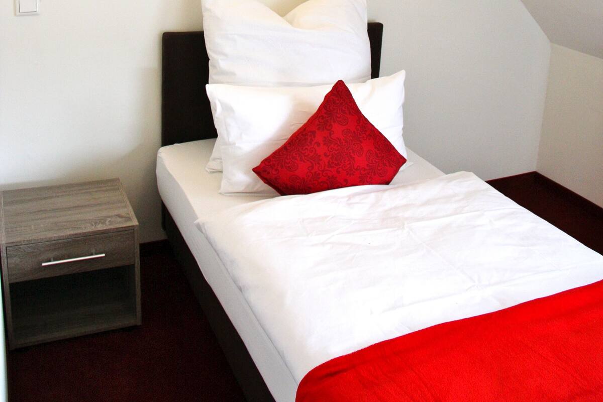 Ein ordentlich bezogenes Bett mit weißer und roter Bettwäsche in einem Zimmer mit schrägem Dach und einer Wandleuchte.