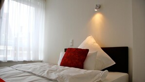 A neatly made bed with white linens and a red pillow, a bedside lamp, and a window with sheer curtains. - Hotel Gemini (Düsseldorf)