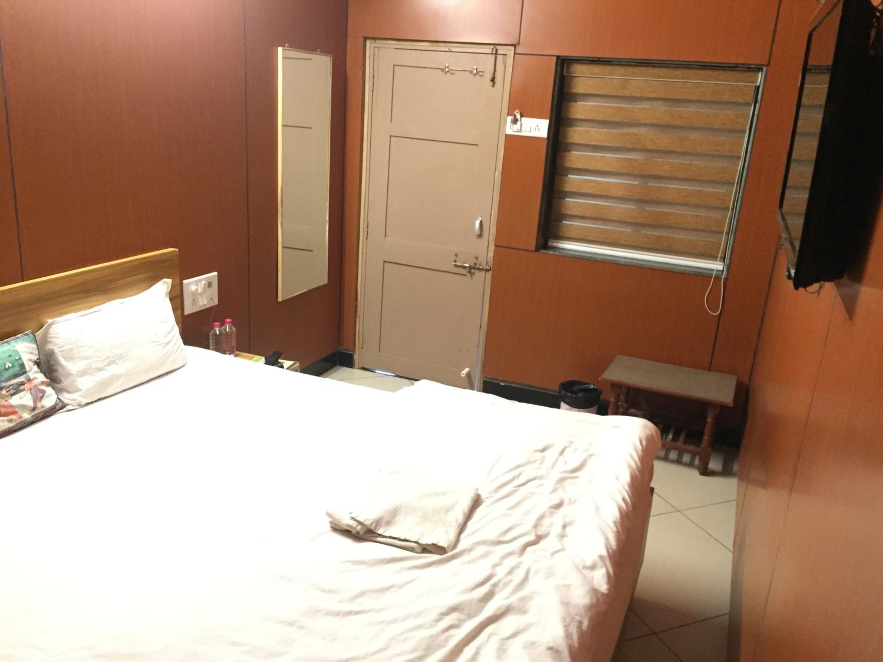 A hotel room with a bed, a bedside table, a mirror, a door, and a window with blinds.