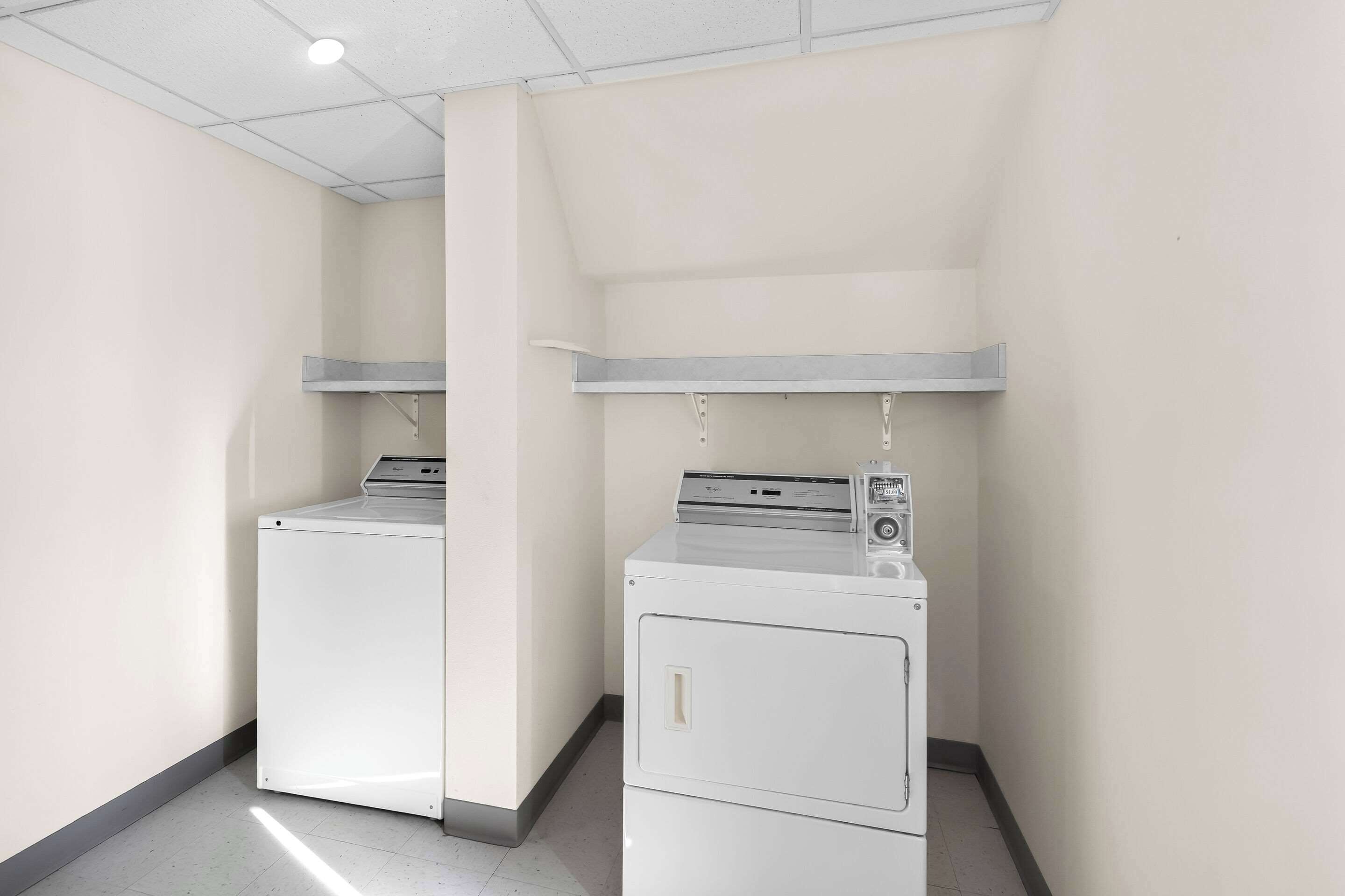 Een wasserette met twee witte wasmachines en drogers, een grijze plank erboven, en een wit plafond met een lamp.