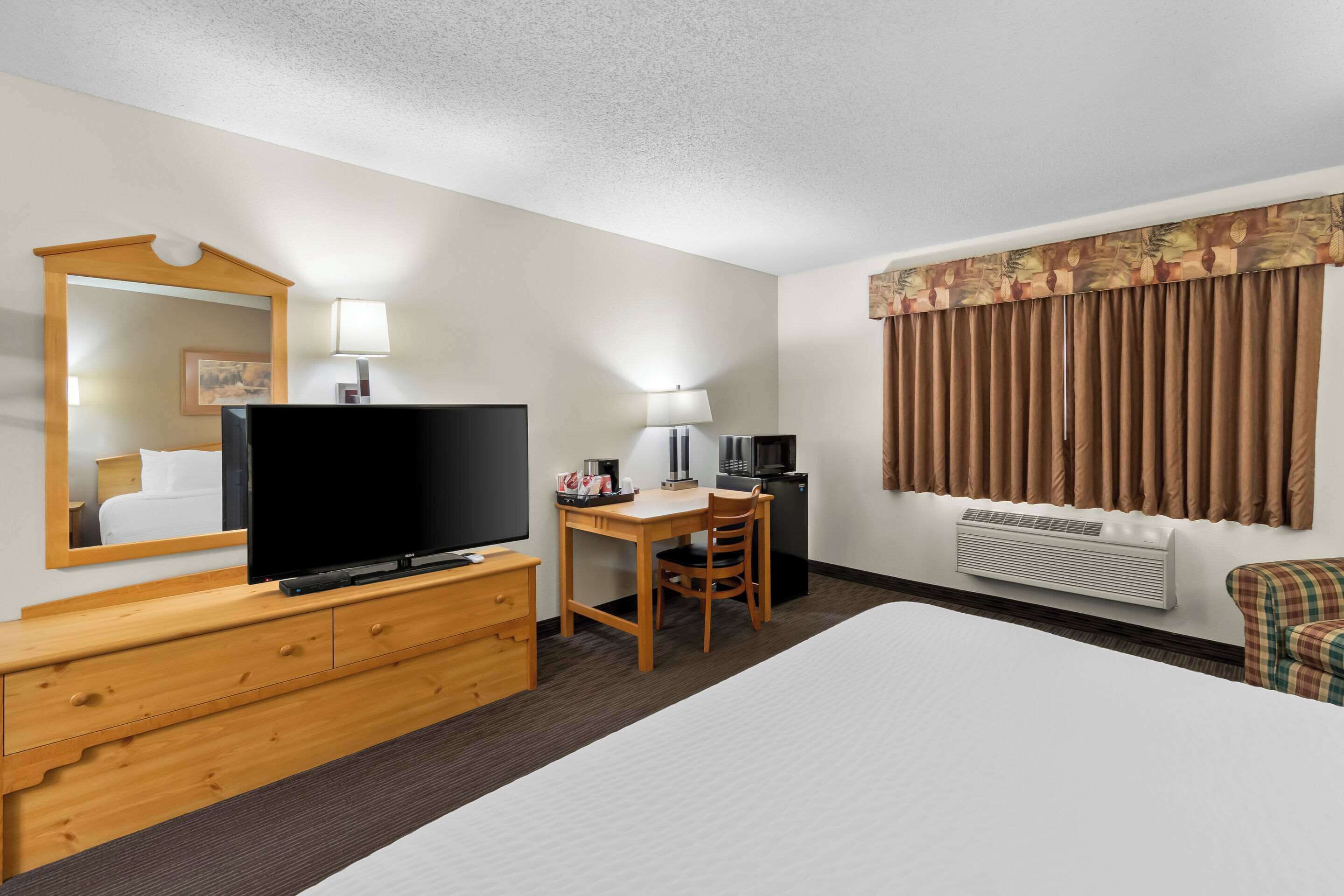 A hotel room with a bed, a desk with a chair, a television, a mirror, a lamp, a heater, and a window with curtains.