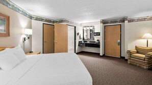 Habitación de hotel con una cama grande, un área de descanso con una silla, un baño con espejo y una kitchenette.