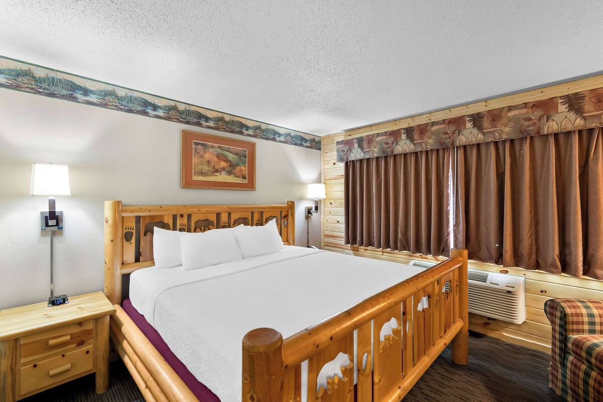 A wooden bed with a headboard, a nightstand with a lamp, a window with curtains, and a painting on the wall.