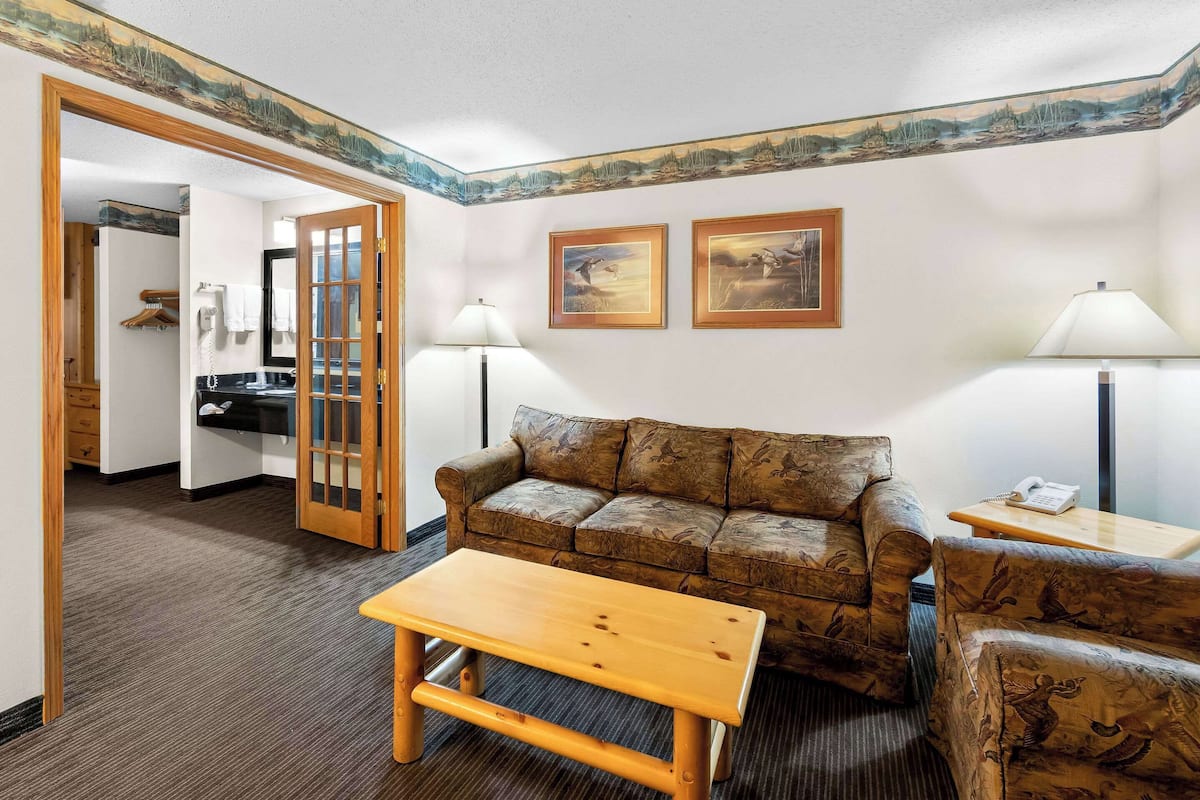 A room with a wooden bench, a patterned sofa, and two lamps. There is a door leading to a bathroom and a hallway.