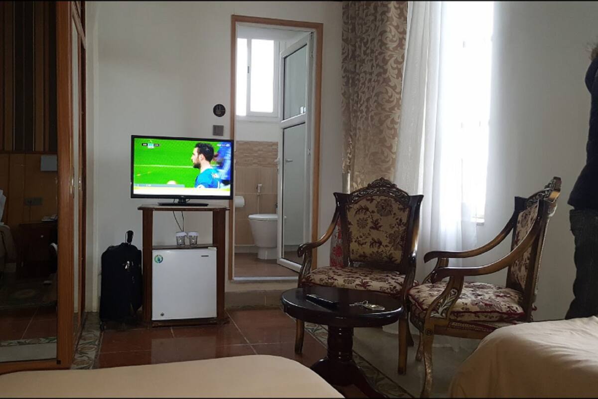 A hotel room with a television displaying a sports game, a small refrigerator, a wooden chair, and a bed.