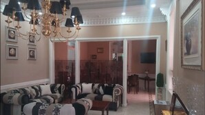 Een woonkamer met een hoekbank, een salontafel en een kroonluchter. Er hangen fotolijstjes aan de muren en er is een eethoek op de achtergrond.