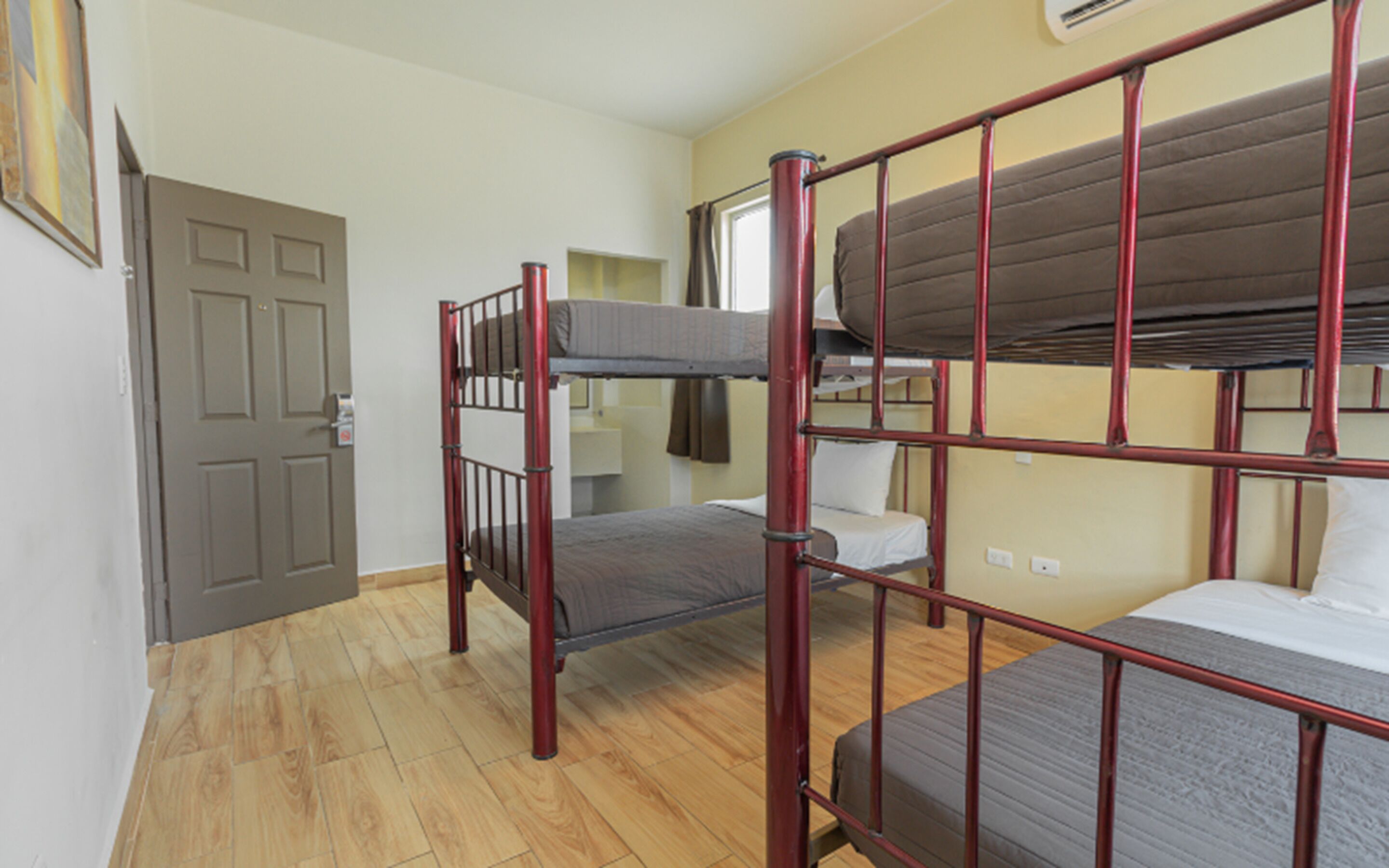 A dormitory room with bunk beds, a wooden floor, a door, and a window.