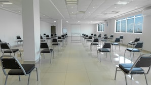 A spacious, well-lit conference room with rows of chairs facing a presentation screen, large windows with a city view, and a tiled floor. - Hotel Plaza del Arco Express (Monterrey)