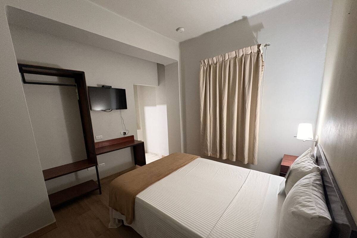 Habitación de hotel con cama, escritorio, televisor, ventana con cortinas y lámpara de noche.